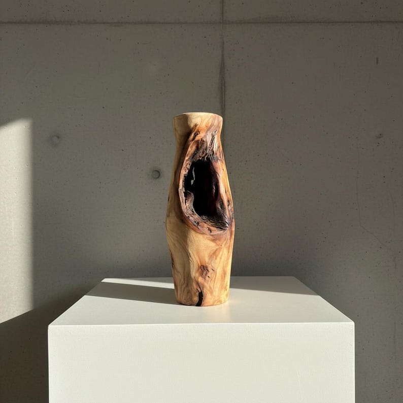 May include: A handcrafted wooden vase with a natural, organic shape. The vase is made from light brown wood and features a large, dark opening in the center. It stands on a white pedestal against a gray concrete wall.