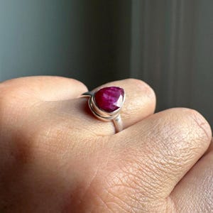 以下が含まれることがあります： 滑らかな金属製のベゼルにセットされた、ファセットカットの洋ナシ型をした深い赤色の宝石が特徴のシルバーリング。石には目に見える内部の質感と光沢のある仕上げが施されており、指を囲むシンプルな丸みを帯びたシルバーバンドに取り付けられています。