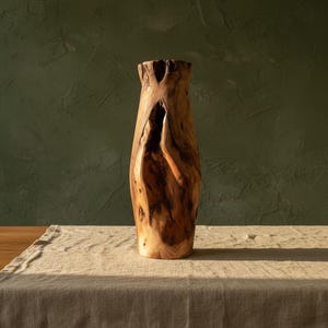 May include: A tall, handcrafted wooden vase with a natural, organic shape. The vase is made from light and dark brown wood, with a rough, textured exterior and a unique, open design. The vase is approximately 12 inches tall.