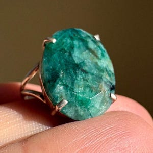 May include: A ring featuring a large, oval-shaped, faceted green gemstone with visible internal inclusions and dark veining. The stone is secured in a silver-toned metal setting with four prongs and a split band design.