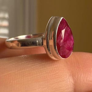 以下が含まれることがあります： 滑らかな金属製のベゼルにセットされた、ファセットカットの洋ナシ型で濃いピンク色の宝石が付いたシルバーリング。バンドは磨かれたシルバーで、石は光を反射するテクスチャーのある多面的な表面をしています。
