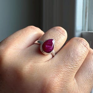 以下が含まれることがあります： 滑らかな金属製のベゼルにセットされた、ティアドロップ型の深いピンクがかった赤色の宝石が特徴のシルバーリング。石の表面はファセットカットされており、指を囲むシンプルで光沢のあるシルバーのバンドに取り付けられています。