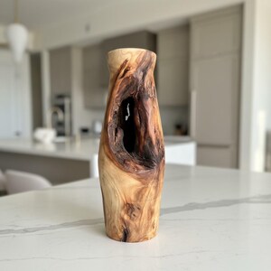 May include: A handcrafted wooden vase with a natural, organic design. The vase features a light brown exterior with dark brown accents and a large, open cavity. The vase is approximately 12 inches tall and has a unique, textured appearance.