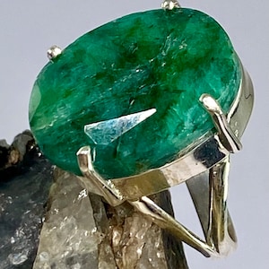 May include: A silver ring featuring a large, oval emerald gemstone. The emerald is a deep green colour and has a faceted cut. The ring is set in a simple, modern style.