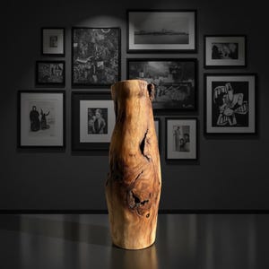 May include: A tall, natural wood vase with a rough, textured surface. The vase is a light brown color with darker brown knots and imperfections. The vase is displayed in front of a gallery wall with framed black and white art.