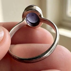 May include: A silver ring featuring a smooth, oval-shaped purple cabochon gemstone set in a simple metal bezel. The band is thin and polished, showing a metallic sheen. The setting holds the stone securely, displaying a light purple hue.