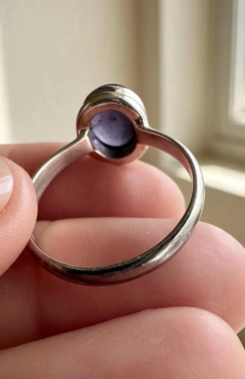 May include: A silver ring featuring a smooth, oval-shaped purple cabochon gemstone set in a simple metal bezel. The band is thin and polished, showing a metallic sheen. The setting holds the stone securely, displaying a light purple hue.