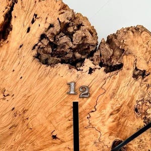 May include: A wooden clock face with a natural, textured grain. The clock features a silver numeral 12 and black clock hands. The wood has a light brown colour with darker brown and black accents.