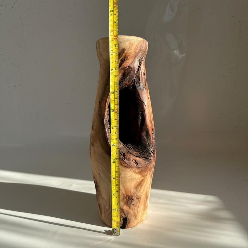 May include: A tall, natural wood vase with a large, irregular opening. The vase is approximately 14 inches tall, with a light brown exterior and darker brown interior. A yellow measuring tape is placed next to the vase.