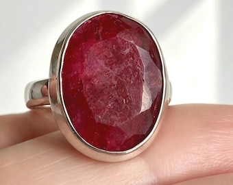 May include: A silver ring featuring a large, oval-shaped, faceted deep red gemstone set in a smooth bezel. The stone displays a textured, translucent surface with natural inclusions, held by a polished silver band.