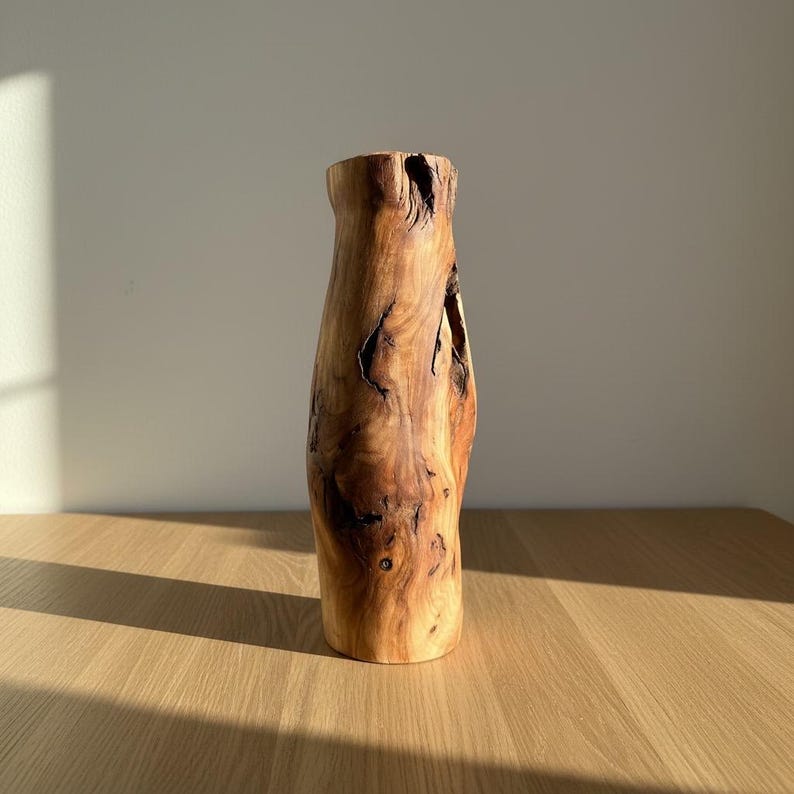 May include: A handcrafted wooden vase with a natural, organic shape. The vase is made from a light brown wood with dark brown knots and grain patterns. The vase is approximately 12 inches tall and has a wide base that tapers to a narrower opening.