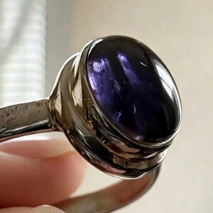 May include: A silver ring featuring a large, oval-shaped, deep purple cabochon gemstone set in a smooth, polished metal bezel. The band is made of a reflective silver-toned metal, and the stone displays a translucent, dark violet hue with natural internal inclusions visible under the light.
