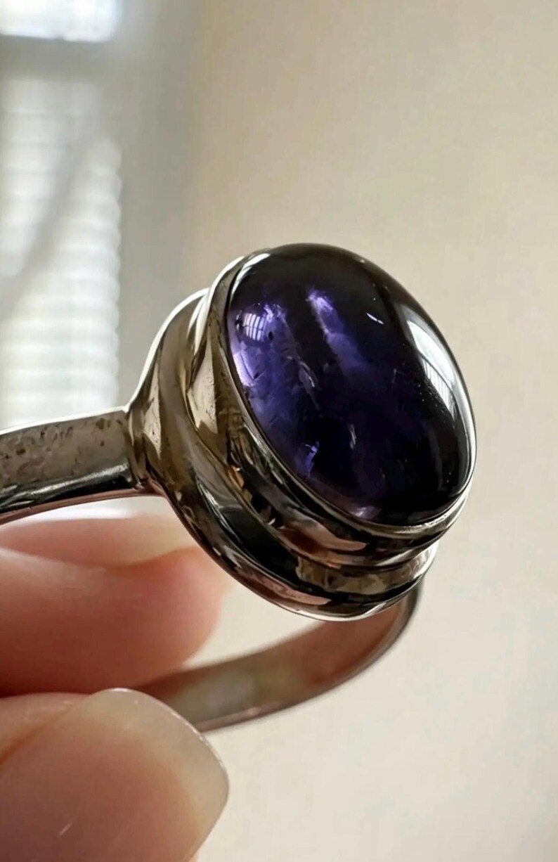 May include: A silver ring featuring a large, oval-shaped, deep purple cabochon gemstone set in a smooth, polished metal bezel. The band is made of a reflective silver-toned metal, and the stone displays a translucent, dark violet hue with natural internal inclusions visible under the light.