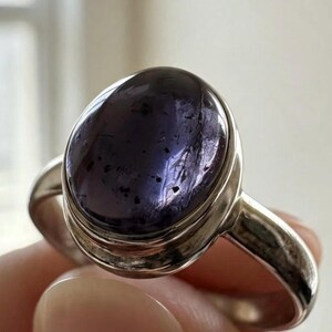 May include: A silver ring featuring a large, oval-shaped, deep purple cabochon gemstone. The stone displays a smooth, polished surface with visible dark inclusions and internal texture. The metal band is plain, polished, and rounded in design.