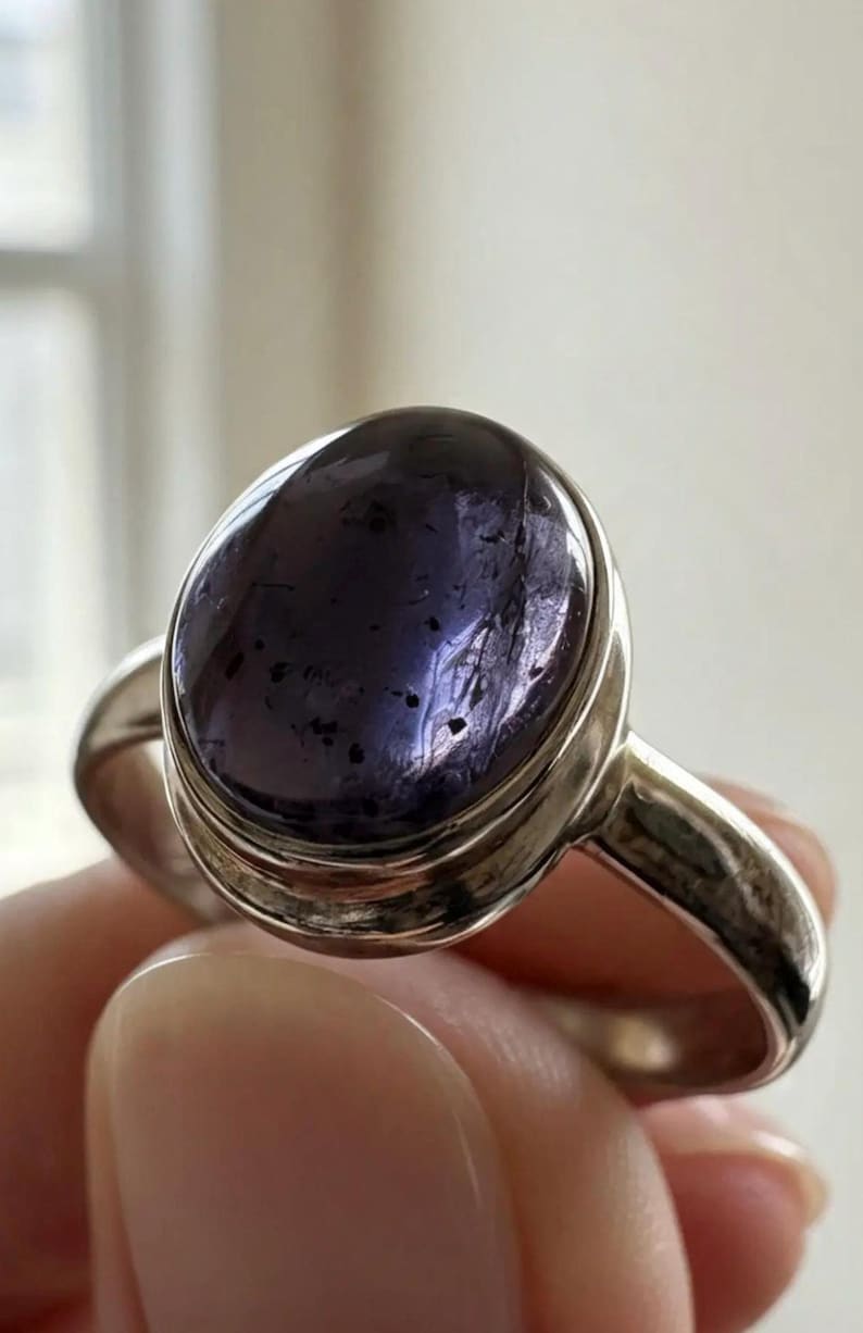 May include: A silver ring featuring a large, oval-shaped, deep purple cabochon gemstone. The stone displays a smooth, polished surface with visible dark inclusions and internal texture. The metal band is plain, polished, and rounded in design.