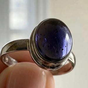 May include: A silver ring featuring a large, oval-shaped, deep purple cabochon gemstone. The stone has a smooth, polished surface with visible dark inclusions and internal fractures. The metal band is plain, polished, and has a simple bezel setting.