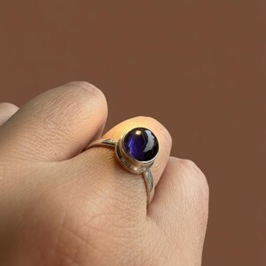 May include: A silver ring featuring a round, polished, deep purple cabochon gemstone set in a smooth metal bezel. The band is thin and metallic, showcasing a reflective surface on the stone that catches the light, highlighting its dark violet hue.