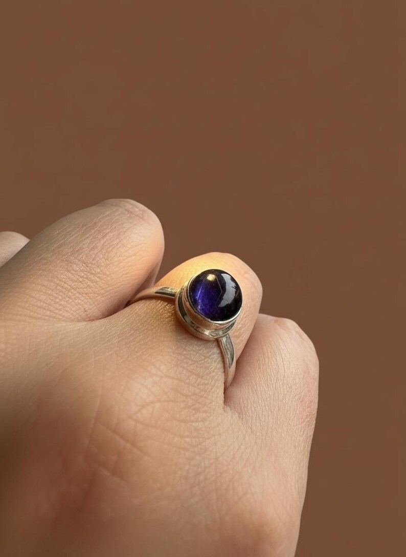 May include: A silver ring featuring a round, polished, deep purple cabochon gemstone set in a smooth metal bezel. The band is thin and metallic, showcasing a reflective surface on the stone that catches the light, highlighting its dark violet hue.