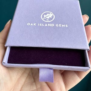 May include: A square, light purple cardboard jewellery box with a sliding drawer mechanism. The top features a white circular logo with an oak leaf and the text OAK ISLAND GEMS. The interior of the drawer is lined with a dark purple velvet fabric.