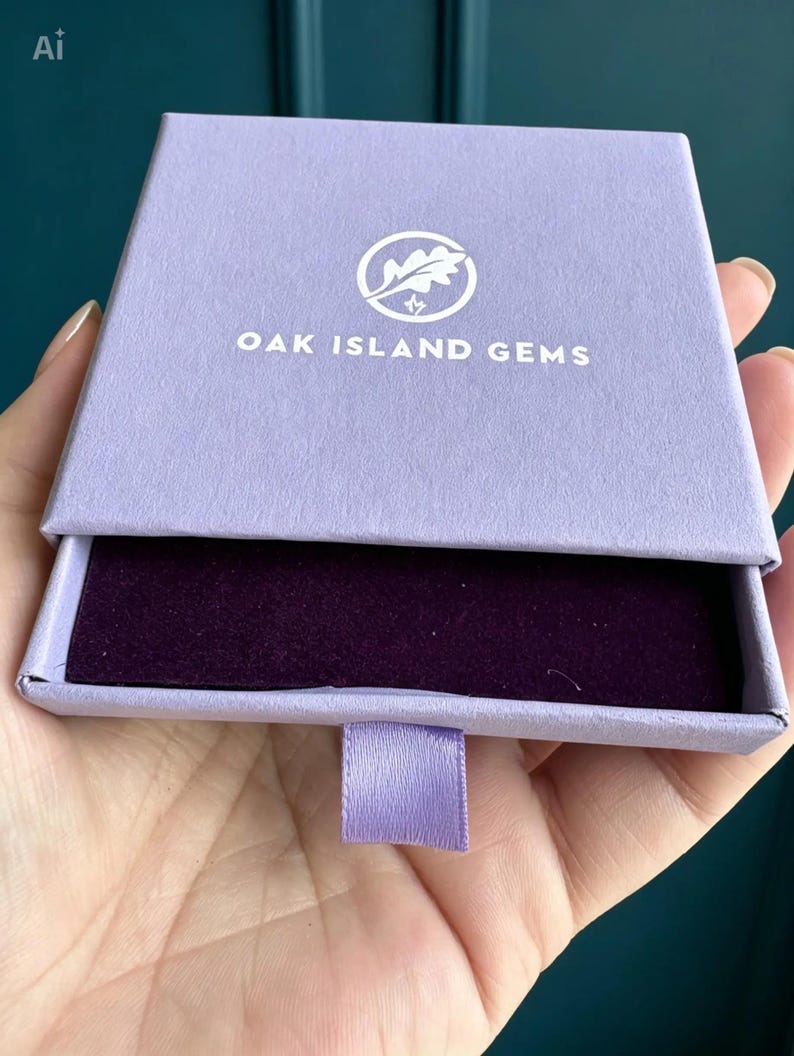 May include: A square, light purple cardboard jewellery box with a sliding drawer mechanism. The top features a white circular logo with an oak leaf and the text OAK ISLAND GEMS. The interior of the drawer is lined with a dark purple velvet fabric.