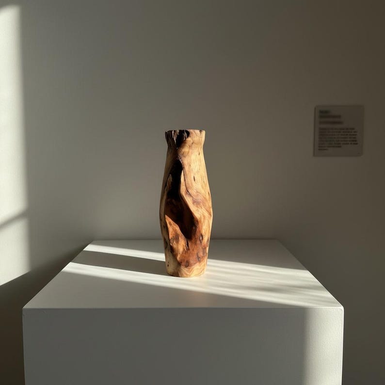 May include: A tall, handcrafted wooden vase with a natural, organic shape. The vase is made from light and dark brown wood, with a textured surface and a flared top. It stands on a white pedestal, illuminated by sunlight.