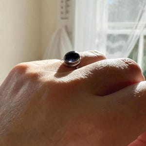 May include: A silver ring featuring a smooth, oval-shaped dark purple gemstone set in a polished metal band. The stone has a deep, reflective surface that catches the light, and the band is simple and rounded, encircling the finger securely.