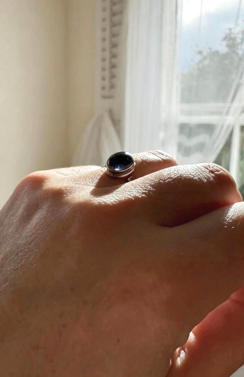 May include: A silver ring featuring a smooth, oval-shaped dark purple gemstone set in a polished metal band. The stone has a deep, reflective surface that catches the light, and the band is simple and rounded, encircling the finger securely.