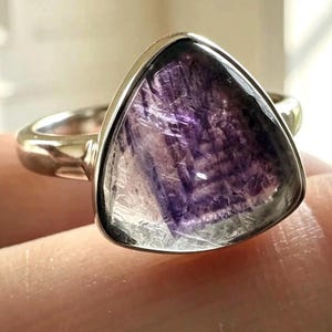 May include: A silver ring featuring a triangular-shaped stone with a translucent appearance and deep purple, geometric inclusions. The metal band is smooth and polished, holding the stone in a simple, secure bezel setting that highlights the gem.