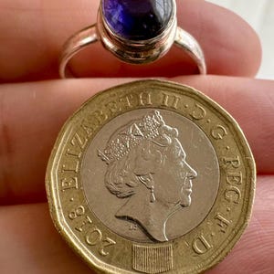 May include: A silver ring featuring a smooth, round, deep purple cabochon gemstone. The ring is positioned directly above a British one-pound coin, which displays the profile of Queen Elizabeth II and the year 2018, for size comparison.