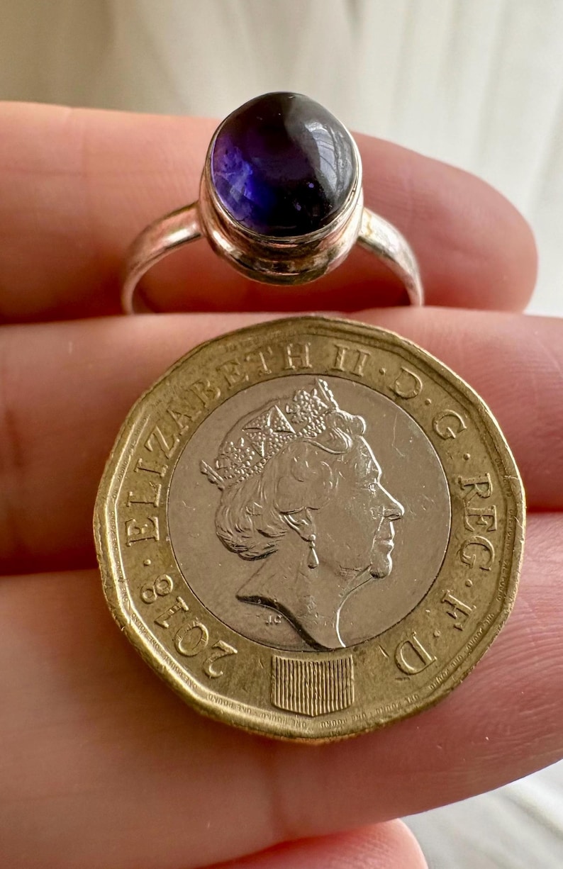 May include: A silver ring featuring a smooth, round, deep purple cabochon gemstone. The ring is positioned directly above a British one-pound coin, which displays the profile of Queen Elizabeth II and the year 2018, for size comparison.