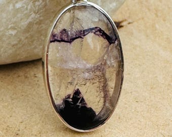 May include: Oval pendant with a clear, textured stone featuring purple and black inclusions, set in a silver-colored metal frame. The pendant hangs from a silver chain, showcasing a natural gemstone design.