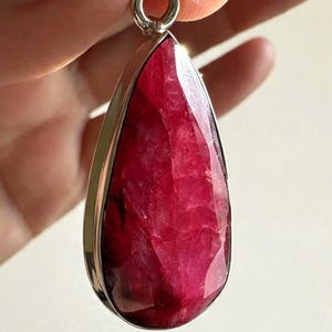 May include: A teardrop-shaped pendant featuring a faceted, deep magenta-red gemstone set in a polished silver-toned metal bezel. The stone displays a textured, crystalline surface with natural inclusions and a pointed top bail for hanging.