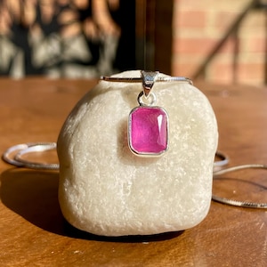 May include: A silver necklace featuring a rectangular, pink gemstone pendant. The pendant is set in a silver bezel and hangs on a delicate silver chain. The necklace is displayed on a white stone. The background is blurred.