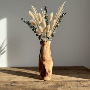 May include: A wooden vase with a natural, organic shape, filled with dried bunny tail grass and eucalyptus. The vase has a warm, brown color and sits on a wooden table. The arrangement is a decorative home accent.