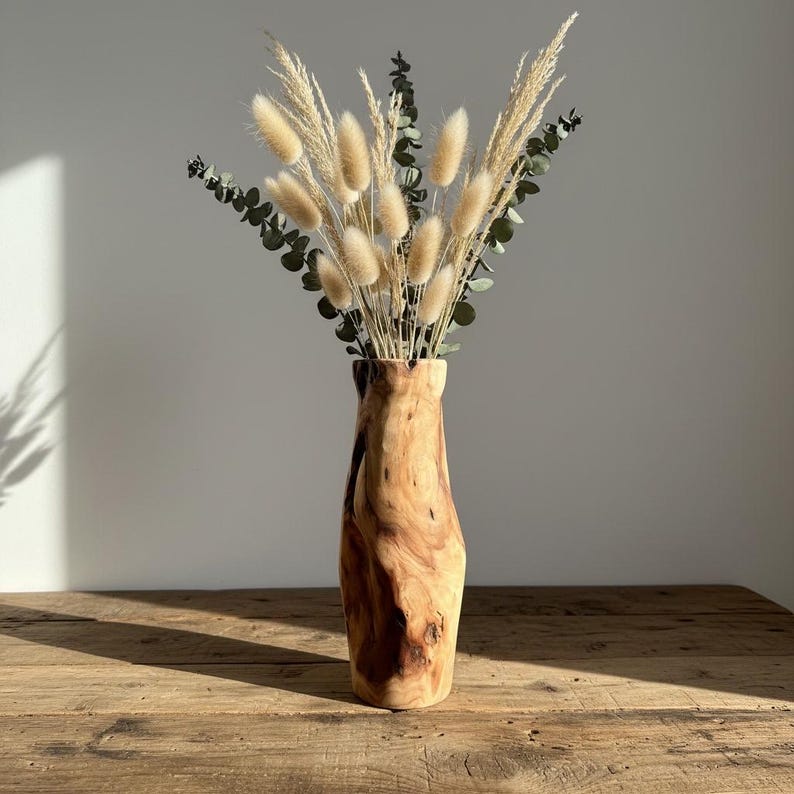 May include: A wooden vase with a natural, organic shape, filled with dried bunny tail grass and eucalyptus. The vase has a warm, brown color and sits on a wooden table. The arrangement is a decorative home accent.