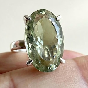 May include: A silver ring featuring a large, oval-cut, light green gemstone. The stone is faceted and held in place by four polished silver prongs. The band of the ring is smooth, solid silver, providing a clean and simple setting for the stone.