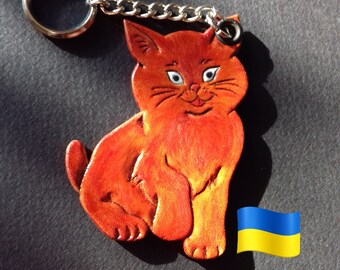 May include: Orange leather keychain shaped like a cat with white eyes and a black nose. The cat is sitting with its tail curled around its legs. The keychain has a silver ring and chain.