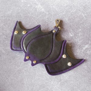May include: A black leather bat-shaped keychain with purple trim and gold-colored rivets. The keychain has a gold-colored clasp for attaching to keys or a bag. The design is stylized and decorative.