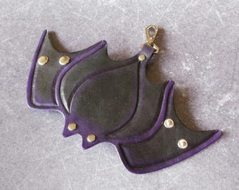 May include: A black leather bat-shaped keychain with purple trim and gold-colored rivets. The keychain has a gold-colored clasp for attaching to keys or a bag. The design is stylized and decorative.