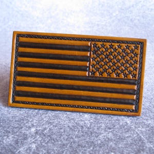 Puede incluir: Un parche rectangular de cuero con un diseño en relieve de la bandera estadounidense. La bandera muestra franjas horizontales alternas en color tostado y marrón oscuro con un campo de estrellas en la esquina superior izquierda, bordeado.