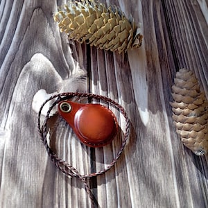 Op de afbeelding: Een bruin leren kompas met een gevlochten koord, op een houten achtergrond met dennenappels.