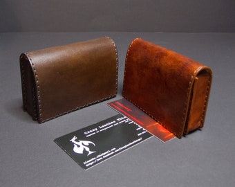May include: Two leather business card holders in brown and reddish-brown, with visible stitching. A business card with a logo and text is in front of the holders. The holders are rectangular with rounded edges.