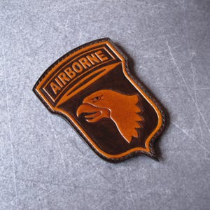 Pode incluir: Um remendo de couro marrom com as palavras "AIRBORNE" em um contorno branco e uma cabeça de águia marrom com um olho branco. O remendo é costurado com fio marrom.