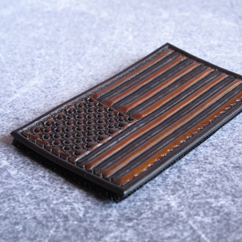 May include: A rectangular patch featuring a debossed American flag design in shades of dark brown and black. The flag pattern includes a textured canton with stars and horizontal stripes, finished with a dark border and a black backing layer.