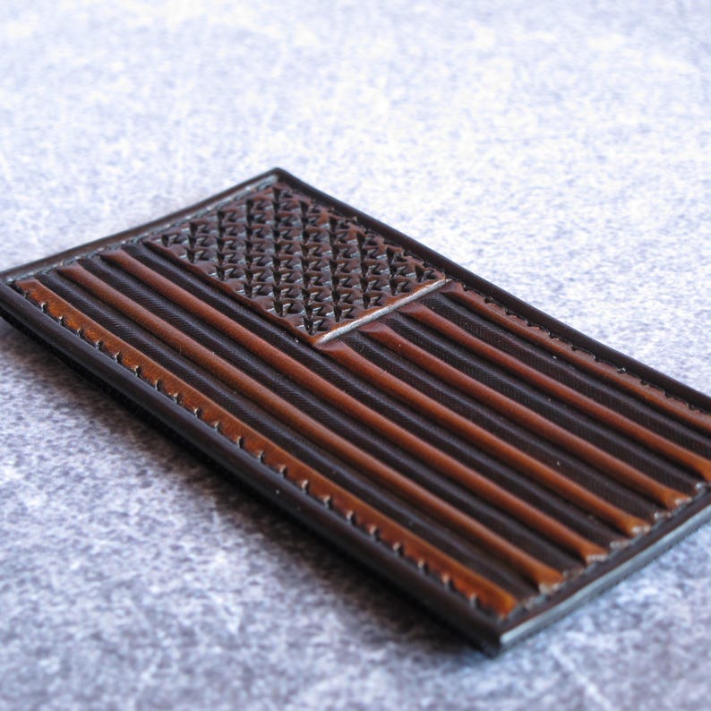 May include: A rectangular leather patch featuring a debossed American flag design. The flag consists of a textured canton with star-like impressions and horizontal stripes in alternating shades of dark brown and a lighter, reddish-brown tone.