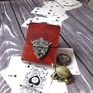 Può includere: Un porta carte in pelle marrone con una chiusura argentata e un design decorato. Il porta carte è su una superficie di legno con un mazzo di carte da gioco e una piccola statuetta di tartaruga dorata.