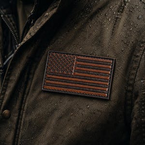 May include: A dark olive green waxed jacket featuring a rectangular leather patch embossed with the American flag. The jacket material has a visible water-resistant texture with small droplets of water scattered across the surface of the fabric.