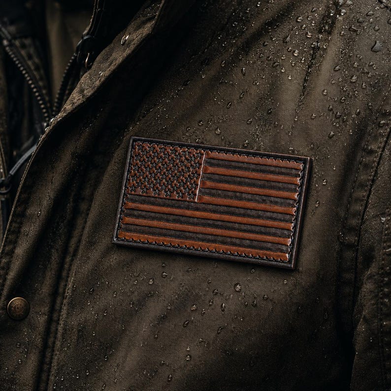 May include: A dark olive green waxed jacket featuring a rectangular leather patch embossed with the American flag. The jacket material has a visible water-resistant texture with small droplets of water scattered across the surface of the fabric.