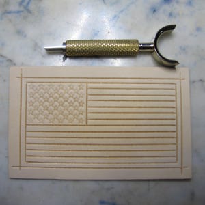 May include: A rectangular piece of light beige vegetable-tanned leather featuring a stamped American flag design with stars and stripes, positioned below a brass-colored metal swivel knife with a textured grip and a curved yoke handle.