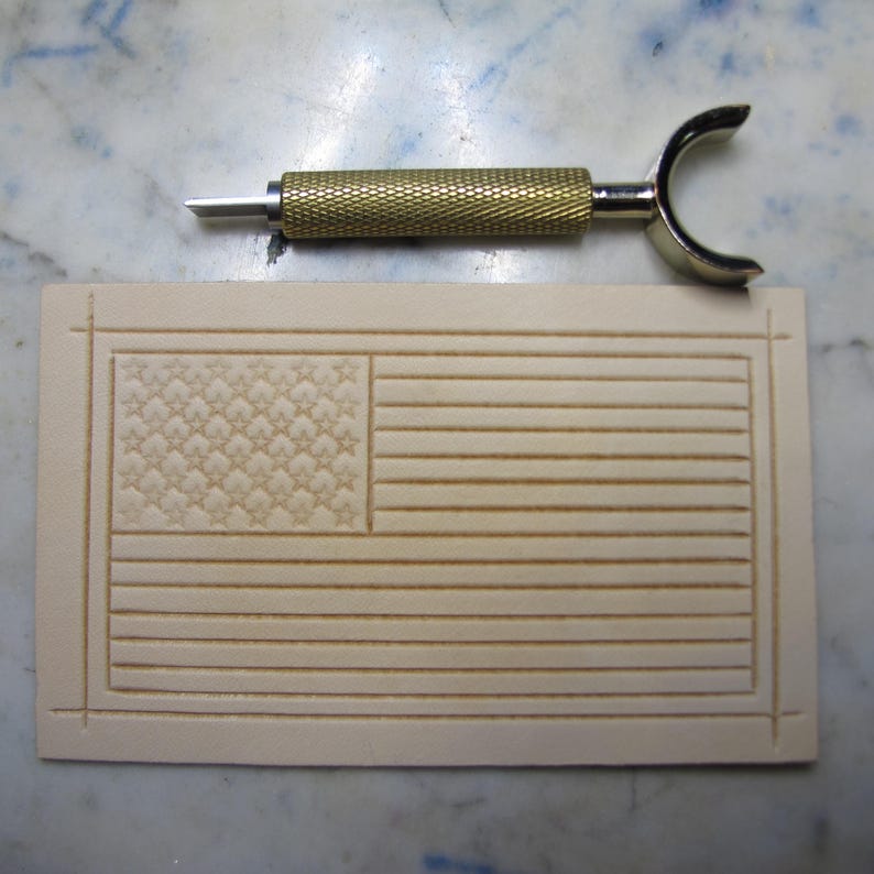 May include: A rectangular piece of light beige vegetable-tanned leather featuring a stamped American flag design with stars and stripes, positioned below a brass-colored metal swivel knife with a textured grip and a curved yoke handle.